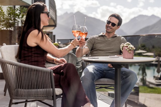 Ein Paar prostet sich mit Aperol Spritz auf einer Terrasse zu, Berge im Hintergrund.