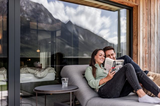 Ein Paar liest gemeinsam ein Buch auf einer Sonnenliege vor Bergaussicht.