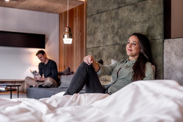 Ein Paar entspannt sich in einem stilvollen Hotelzimmer, sie auf dem Bett, er liest.