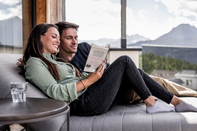 Glückliches Paar entspannt auf einem Sofa, liest ein Buch mit Bergblick.