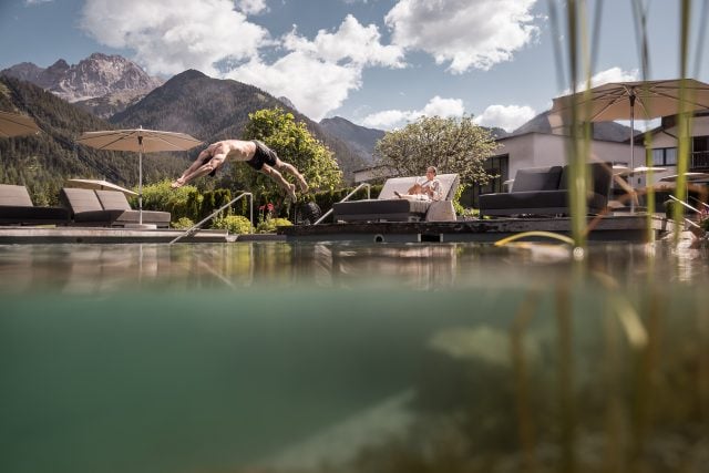 Mann springt in einen Pool vor Bergkulisse, Frau entspannt im Liegestuhl.