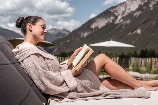 Frau liest Buch im Bademantel auf Liege in den Bergen.