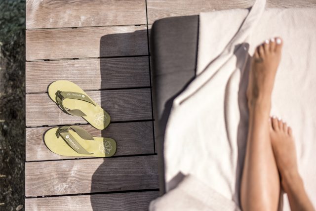 Grüne Flip-Flops auf Holzplanken neben den Füßen auf einem Handtuch.