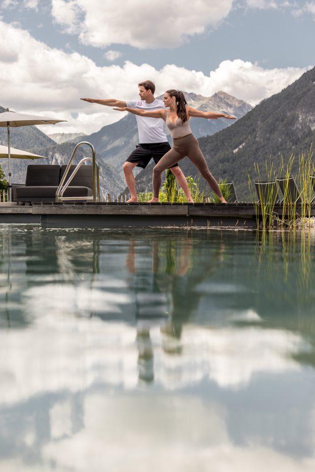 Paar praktiziert Yoga am Pool mit Bergpanorama im Hintergrund.